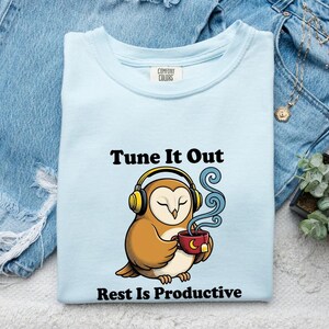 May include: A light blue t-shirt featuring a graphic of a brown owl wearing yellow headphones and holding a red mug with steam rising. The text above reads Tune It Out and the text below reads Rest Is Productive in black lettering.