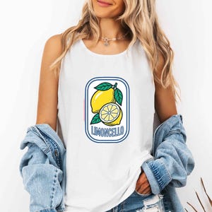 Pode incluir: Uma regata branca sem mangas com uma estampa gráfica de um limão amarelo e uma metade de limão com folhas verdes, contida em uma borda retangular azul com cantos arredondados e o texto LIMONCELLO impresso em letras azuis.