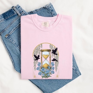 May include: A light pink t-shirt featuring a graphic print of a gold hourglass centered within an arched frame. The design includes three black crows flying around the hourglass and a cluster of blue flowers at the base, all set against blue jeans.