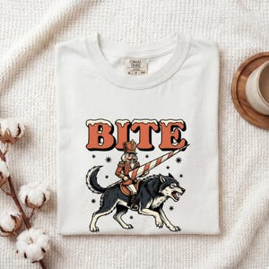 May include: A white t-shirt featuring a graphic of a nutcracker riding a black and grey wolf while holding a candy cane lance. The word BITE is printed in large orange letters with white snow-like accents above the illustration. The shirt has a Comfort Colors brand tag at the neck.