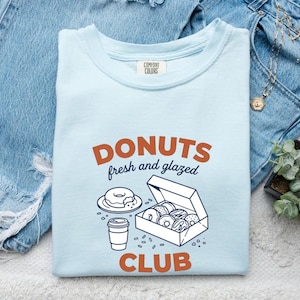 May include: A light blue t-shirt featuring a graphic of a donut on a plate, a coffee cup, and a box of donuts. The text reads "DONUTS fresh and glazed CLUB" in orange and dark blue. The shirt has a Comfort Colors brand label at the neck.