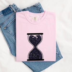 May include: A light pink short-sleeved t-shirt featuring a black graphic of an hourglass filled with multiple black ravens. The shirt has a crew neckline and a Comfort Colors brand label printed on the inside of the collar area.