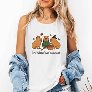 May include: A white sleeveless tank top featuring a graphic of three capybaras, one wearing a pumpkin, one in a green sweater, and one with a witch hat holding an apple basket. The text "Unbothered and autumnal" is printed below the illustration.