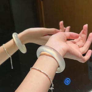 Puede incluir: Un par de brazaletes translúcidos de color claro. Uno se lleva en la muñeca y el otro se refleja en un espejo. También se ve una delicada pulsera de plata. Los brazaletes tienen un diseño suave y redondeado, y la estética general es minimalista y elegante.