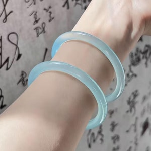 May include: Two light blue jadeite bangles are worn on a person's arm. The translucent bracelets have a smooth, polished finish. The background features blurred calligraphy on a light-colored surface.