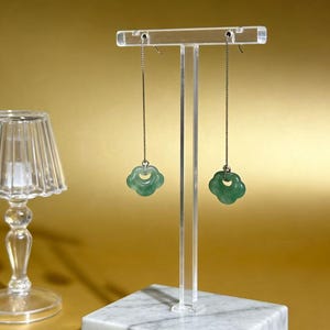 Può includere: Un paio di orecchini di giada con un design a forma di nuvola sono appesi a un espositore trasparente in acrilico. Gli orecchini sono di colore verde chiaro e sono sospesi da delicate catene d'argento. L'espositore poggia su una base quadrata di marmo.