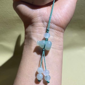 May include: A light green jade-like gourd charm attached to a dark green cord. The charm features a central gourd-shaped bead, one small round bead above it, and two round beads at the bottom ends of the cord, creating a decorative hanging accessory.