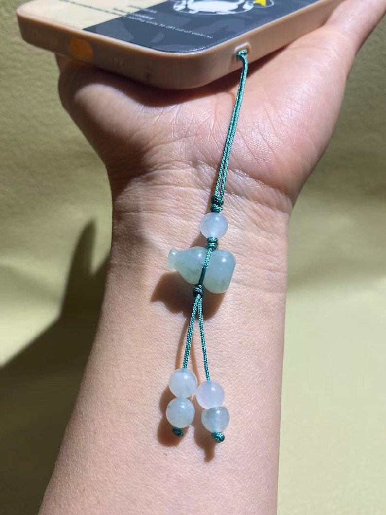 May include: A light green jade-like gourd charm attached to a dark green cord. The charm features a central gourd-shaped bead, one small round bead above it, and two round beads at the bottom ends of the cord, creating a decorative hanging accessory.
