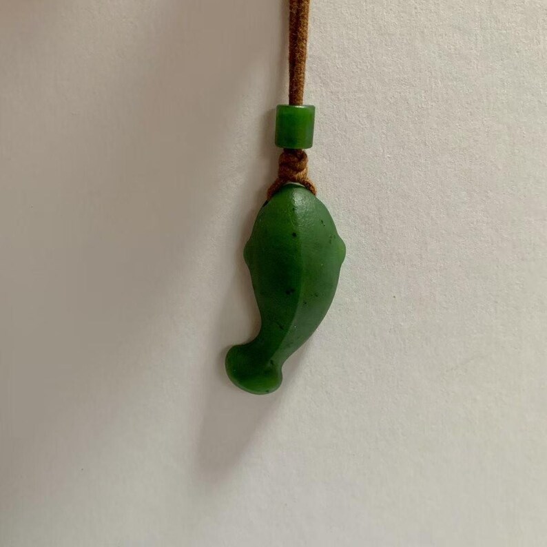 May include: A green jade pendant in the shape of a dolphin, suspended from a brown cord. The dolphin pendant has a smooth, polished surface and is attached to a small green bead. The cord is knotted above the pendant.