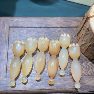 May include: Seven light amber-colored, decorative bottle stoppers. Each stopper has a rounded body, a bulbous base, and a crown-like top. The stoppers are arranged on a wooden surface.
