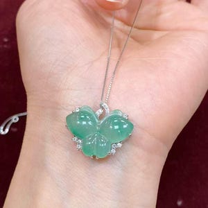 Puede incluir: Colgante de jade verde con forma de mariposa, montado en una delicada cadena de plata. El colgante presenta un color verde claro transl&uacute;cido y un acabado pulido. Peque&ntilde;as piedras preciosas transparentes rodean las alas de la mariposa.