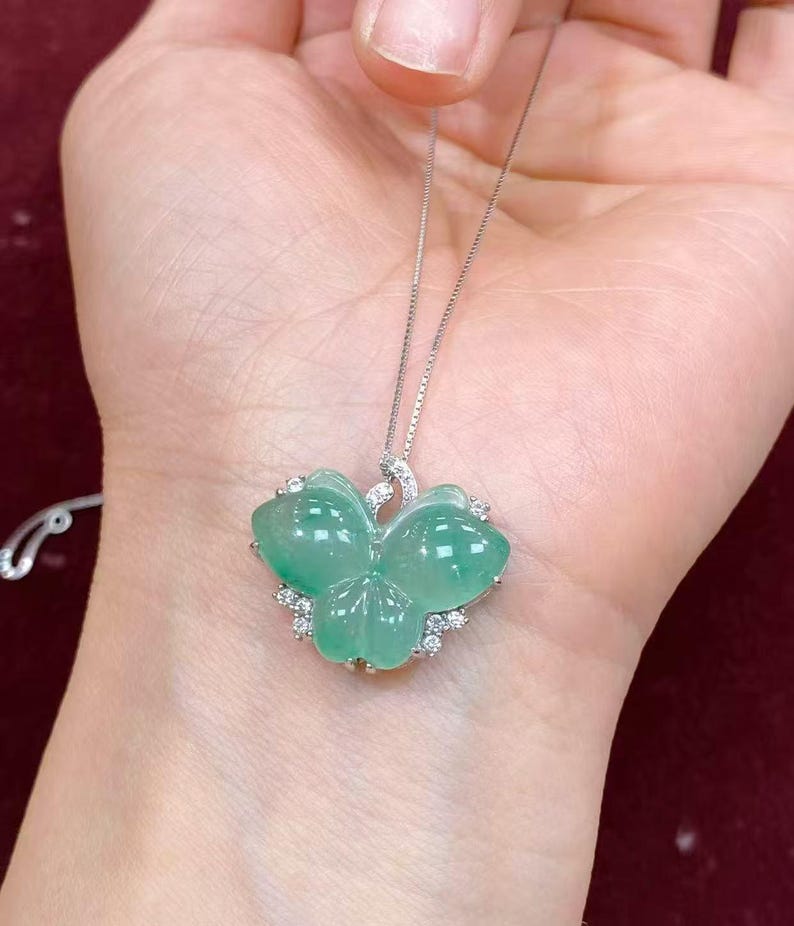 Puede incluir: Colgante de jade verde con forma de mariposa, montado en una delicada cadena de plata. El colgante presenta un color verde claro transl&uacute;cido y un acabado pulido. Peque&ntilde;as piedras preciosas transparentes rodean las alas de la mariposa.
