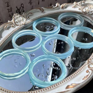May include: A collection of light blue, translucent bangles arranged on a mirrored surface within a decorative, oval-shaped tray. The bangles have a smooth, polished appearance, reflecting light and creating a subtle glow. The tray has a white and gold ornate design.
