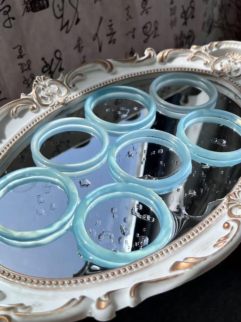 May include: A collection of light blue, translucent bangles arranged on a mirrored surface within a decorative, oval-shaped tray. The bangles have a smooth, polished appearance, reflecting light and creating a subtle glow. The tray has a white and gold ornate design.