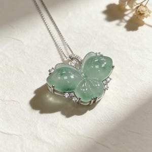 Puede incluir: Un collar de plata con un colgante en forma de mariposa. El colgante est&aacute; hecho de jade verde transl&uacute;cido y est&aacute; adornado con peque&ntilde;as piedras preciosas transparentes. El collar se presenta sobre una superficie blanca, con una delicada cadena.