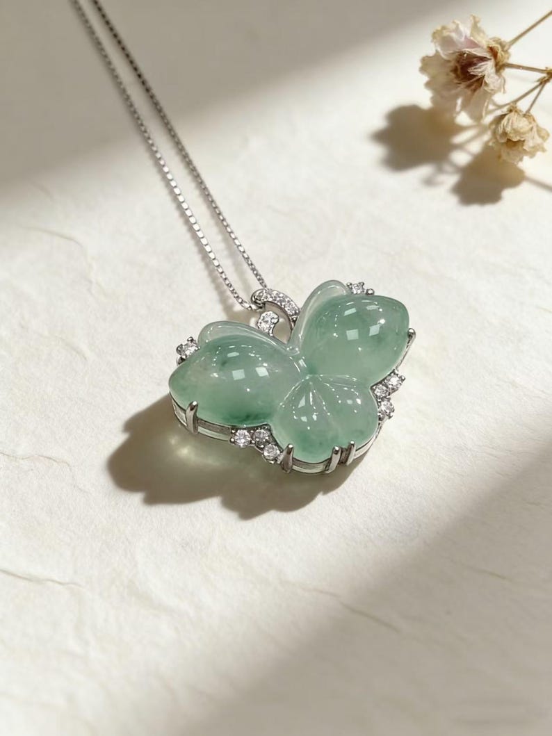 Puede incluir: Un collar de plata con un colgante en forma de mariposa. El colgante est&aacute; hecho de jade verde transl&uacute;cido y est&aacute; adornado con peque&ntilde;as piedras preciosas transparentes. El collar se presenta sobre una superficie blanca, con una delicada cadena.