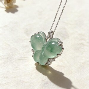 Puede incluir: Un collar con colgante de jade en forma de flor, de color verde claro. El jade est&aacute; engastado en un marco de metal plateado y adornado con peque&ntilde;as piedras preciosas transparentes. El collar cuelga de una delicada cadena plateada.