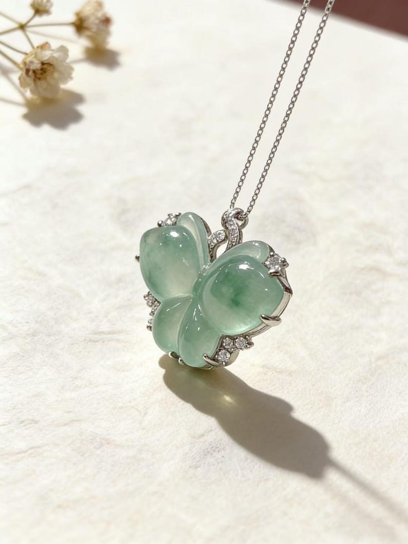 Puede incluir: Un collar con colgante de jade en forma de flor, de color verde claro. El jade est&aacute; engastado en un marco de metal plateado y adornado con peque&ntilde;as piedras preciosas transparentes. El collar cuelga de una delicada cadena plateada.