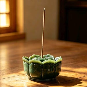 May include: A jade green ceramic lotus flower incense holder with a lit incense stick. The incense holder is glossy and sits on a wooden surface. The incense stick is brown and slender, with a wisp of smoke rising from it.
