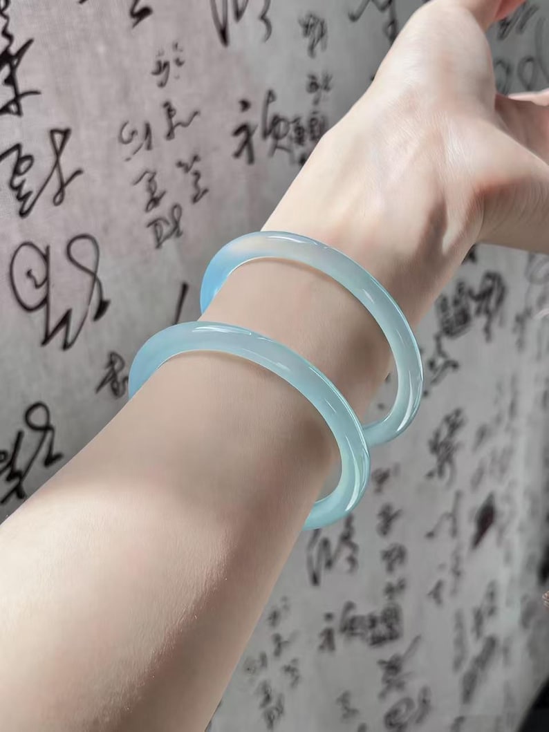 May include: Two light blue jadeite bangles are worn on a person's arm. The translucent bracelets have a smooth, polished finish. The background features blurred calligraphy on a light-colored surface.