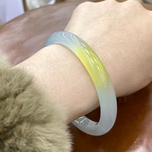 May include: A translucent jade bangle bracelet with a colour gradient, shifting from pale blue to yellow. The smooth, polished surface reflects light, highlighting the natural variations in the stone. This elegant piece is a stylish accessory.