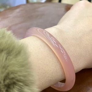 May include: A pale pink bangle bracelet. The smooth, rounded bracelet is worn on a wrist, highlighting its delicate colour and polished surface. The bracelet's soft hue complements the skin tone, creating a subtle, elegant accessory.