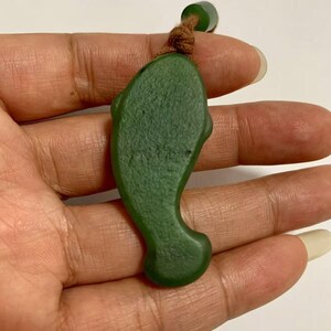 Puede incluir: Un colgante de jade verde con forma de delf&iacute;n. El colgante es de color verde liso y uniforme con un cord&oacute;n marr&oacute;n atado en la parte superior. El colgante de delf&iacute;n se sostiene en una mano.