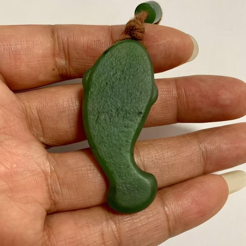 Puede incluir: Un colgante de jade verde con forma de delf&iacute;n. El colgante es de color verde liso y uniforme con un cord&oacute;n marr&oacute;n atado en la parte superior. El colgante de delf&iacute;n se sostiene en una mano.