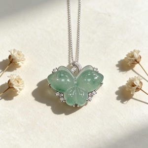 Puede incluir: Un delicado collar de plata con un colgante de mariposa de jade verde claro. El colgante de mariposa est&aacute; adornado con peque&ntilde;as piedras preciosas transparentes en los bordes. El collar se muestra sobre una superficie blanca con peque&ntilde;as flores blancas.