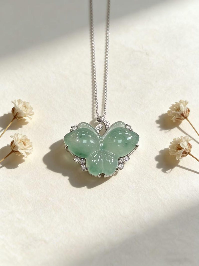 Puede incluir: Un delicado collar de plata con un colgante de mariposa de jade verde claro. El colgante de mariposa est&aacute; adornado con peque&ntilde;as piedras preciosas transparentes en los bordes. El collar se muestra sobre una superficie blanca con peque&ntilde;as flores blancas.