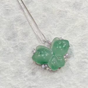 Puede incluir: Un collar de plata con un colgante de jade verde en forma de tr&eacute;bol. El colgante est&aacute; adornado con peque&ntilde;as piedras preciosas transparentes. La cadena es fina y el colgante es el centro de la imagen.