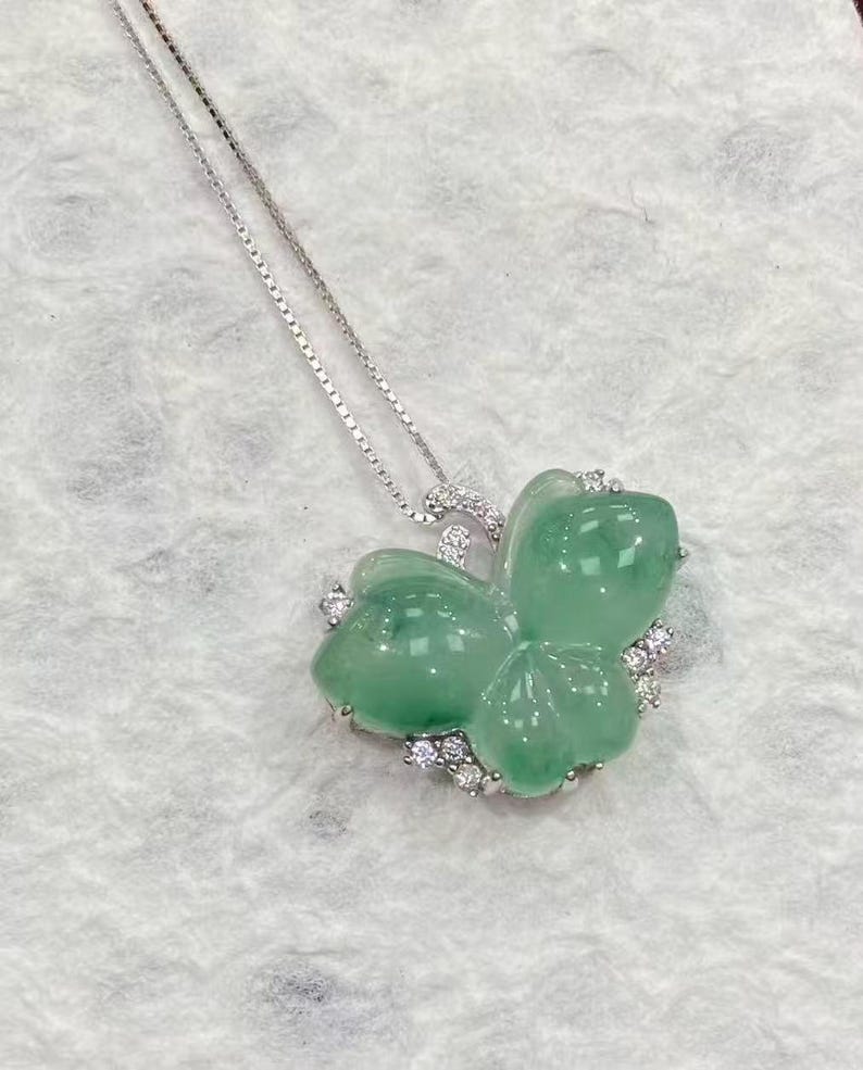 Puede incluir: Un collar de plata con un colgante de jade verde en forma de tr&eacute;bol. El colgante est&aacute; adornado con peque&ntilde;as piedras preciosas transparentes. La cadena es fina y el colgante es el centro de la imagen.