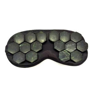 May include: A black eye mask featuring numerous dark green, hexagonal stones. The stones are arranged in two sections, covering the eye area. The mask is designed for relaxation and wellness.
