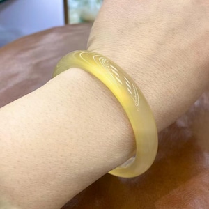 May include: A translucent yellow jade bangle bracelet. The smooth, polished surface of the bracelet is a soft yellow colour. The bracelet has a simple, elegant design, with a subtle etched pattern on the surface.