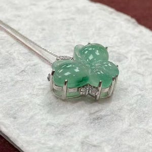 Puede incluir: Un collar de plata con un colgante en forma de tr&eacute;bol de cuatro hojas. El colgante est&aacute; hecho de un material transl&uacute;cido de color verde claro y est&aacute; adornado con peque&ntilde;as piedras transparentes. El collar se muestra sobre una superficie blanca texturizada.
