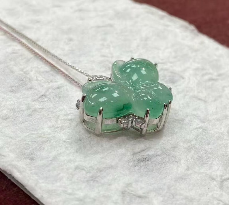Puede incluir: Un collar de plata con un colgante en forma de tr&eacute;bol de cuatro hojas. El colgante est&aacute; hecho de un material transl&uacute;cido de color verde claro y est&aacute; adornado con peque&ntilde;as piedras transparentes. El collar se muestra sobre una superficie blanca texturizada.