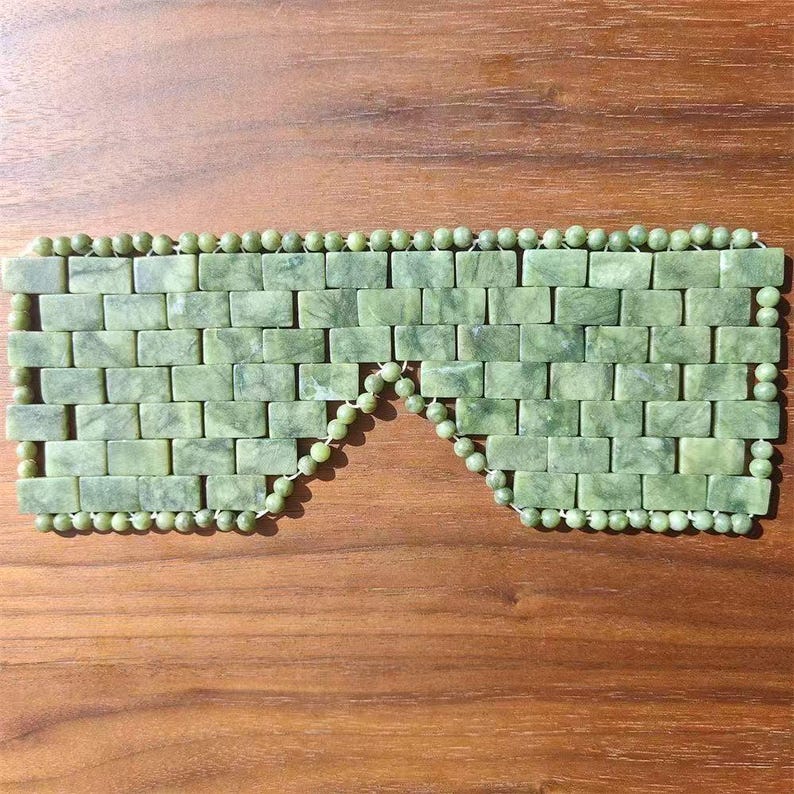 Puede incluir: Una m&aacute;scara de ojos de jade verde con un patr&oacute;n similar a ladrillos. La m&aacute;scara est&aacute; hecha de peque&ntilde;as piedras de jade rectangulares y est&aacute; bordeada con peque&ntilde;as cuentas de jade redondas. Se muestra sobre una superficie de madera.