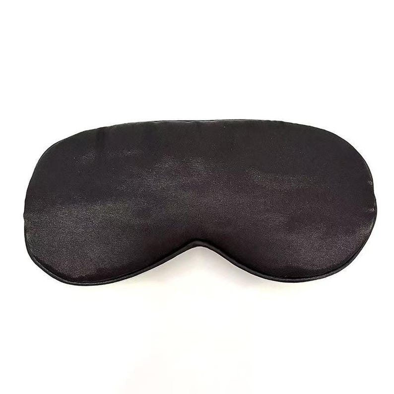 May include: A black sleep mask with a smooth, satin-like texture. The mask is oval-shaped, designed to cover the eyes and block out light. It is a solid, dark colour, and appears to be made of a soft material.