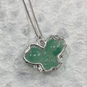 Puede incluir: Un collar de plata con un colgante en forma de hoja verde. El colgante de hoja tiene una superficie lisa y pulida y est&aacute; engastado en un marco plateado con peque&ntilde;os detalles decorativos. La cadena es una fina cadena de plata.
