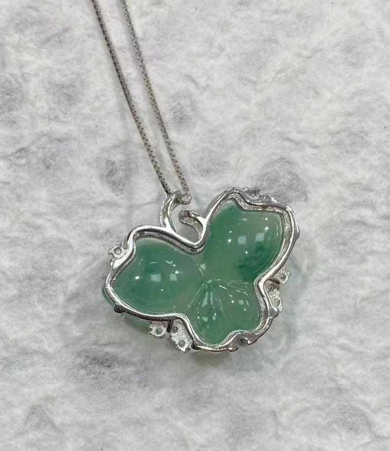 Puede incluir: Un collar de plata con un colgante en forma de hoja verde. El colgante de hoja tiene una superficie lisa y pulida y est&aacute; engastado en un marco plateado con peque&ntilde;os detalles decorativos. La cadena es una fina cadena de plata.
