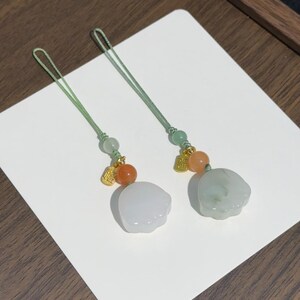 May include: Two decorative charms featuring light green cord loops, small green beads, orange spherical beads, and gold-toned metal charms with Chinese characters. Each charm ends in a smooth, pale, cloud-shaped stone pendant with a polished finish.