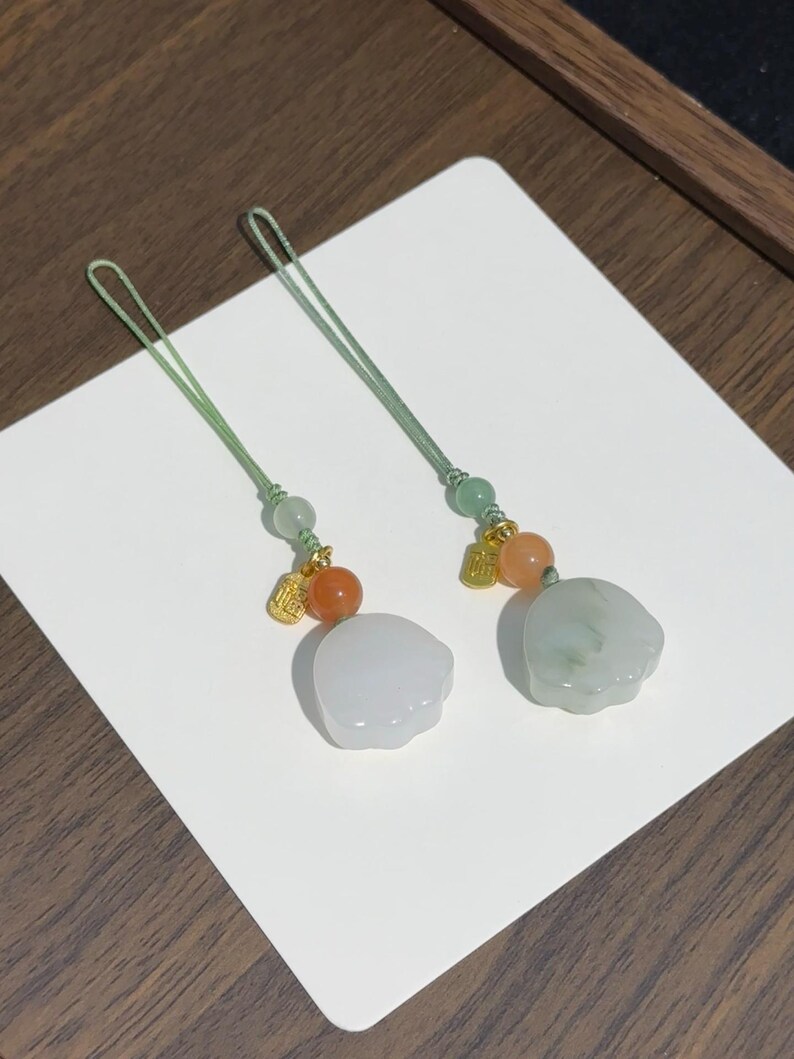May include: Two decorative charms featuring light green cord loops, small green beads, orange spherical beads, and gold-toned metal charms with Chinese characters. Each charm ends in a smooth, pale, cloud-shaped stone pendant with a polished finish.
