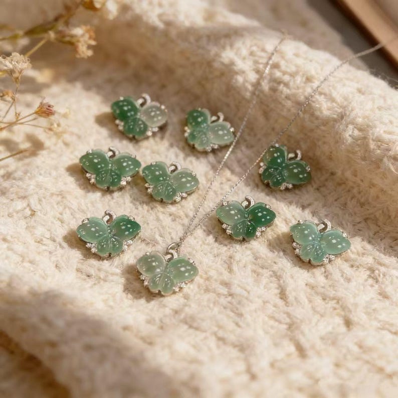 Puede incluir: Una colecci&oacute;n de colgantes y pendientes verdes con forma de mariposa, con detalles blancos y engastes plateados. Un colgante cuelga de una fina cadena de plata. Las joyas se exhiben sobre una superficie texturizada de color crema.
