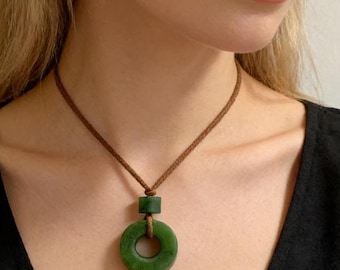 May include: A green jade pendant necklace. The pendant features a circular jade stone with a smaller rectangular jade piece above it, suspended from a brown cord. The necklace is displayed against a neutral background.