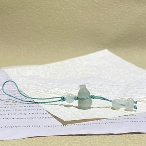 May include: A light green, translucent, gourd-shaped stone charm suspended on a thin, teal-colored cord. The cord features a loop at one end and is accented with several small, matching light green, spherical stone beads knotted along the string.