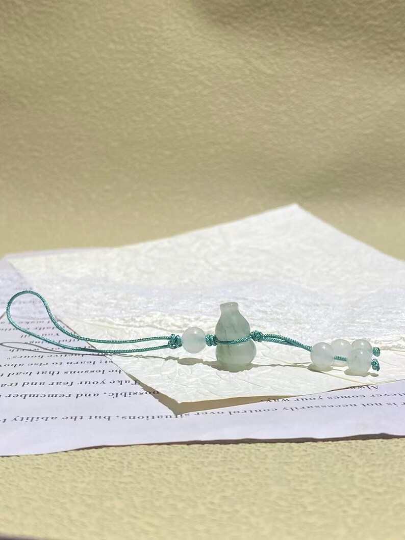 May include: A light green, translucent, gourd-shaped stone charm suspended on a thin, teal-colored cord. The cord features a loop at one end and is accented with several small, matching light green, spherical stone beads knotted along the string.