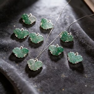 Puede incluir: Una colecci&oacute;n de colgantes y collares con forma de mariposa. Los colgantes son de color verde transl&uacute;cido con motas blancas y est&aacute;n engarzados en plata con peque&ntilde;as piedras preciosas transparentes. Las joyas se exhiben sobre una superficie aterciopelada de color gris oscuro.