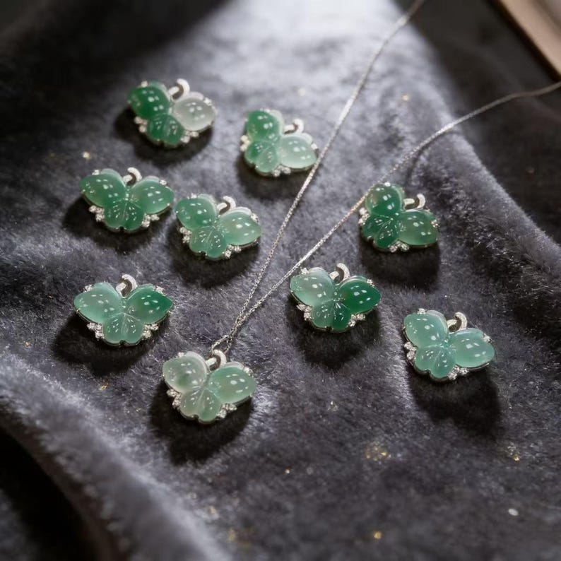 Puede incluir: Una colecci&oacute;n de colgantes y collares con forma de mariposa. Los colgantes son de color verde transl&uacute;cido con motas blancas y est&aacute;n engarzados en plata con peque&ntilde;as piedras preciosas transparentes. Las joyas se exhiben sobre una superficie aterciopelada de color gris oscuro.