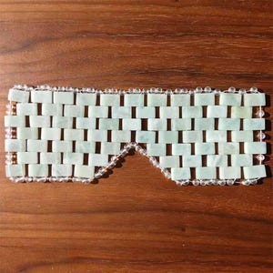 Puede incluir: Una m&aacute;scara de ojos de jade con un patr&oacute;n de ladrillos. La m&aacute;scara est&aacute; hecha de piedras de jade rectangulares de color verde claro, conectadas por cuentas transparentes. La m&aacute;scara est&aacute; dise&ntilde;ada para cubrir los ojos y se muestra sobre una superficie de madera.