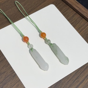 May include: Two decorative charms featuring cylindrical, pale green stone pendants suspended from light green cords. Each charm includes a small, translucent white bead, a vibrant orange bead, and a tiny gold-toned spacer accenting the cord.